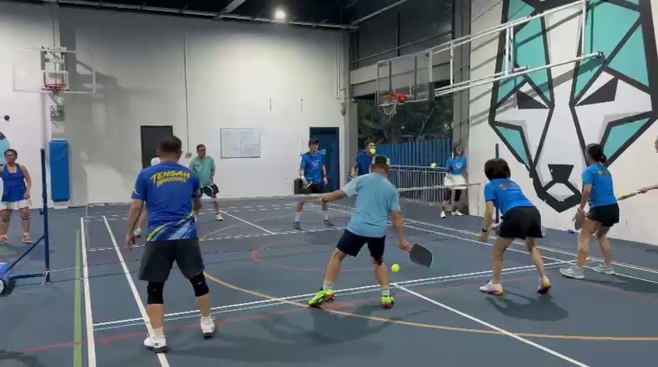 Players in club shirts warm up on an indoor court, rallying a pickleball across the net before the first serve of opening night.